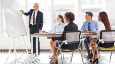 Positive senior leader standing at flipchart at meeting at drawing graph to his team. Multiethnic business people looking at him and smiling