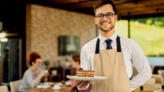 Happy waiter with slice of cake standing in a cafe and looking at camera. There are people in the background.